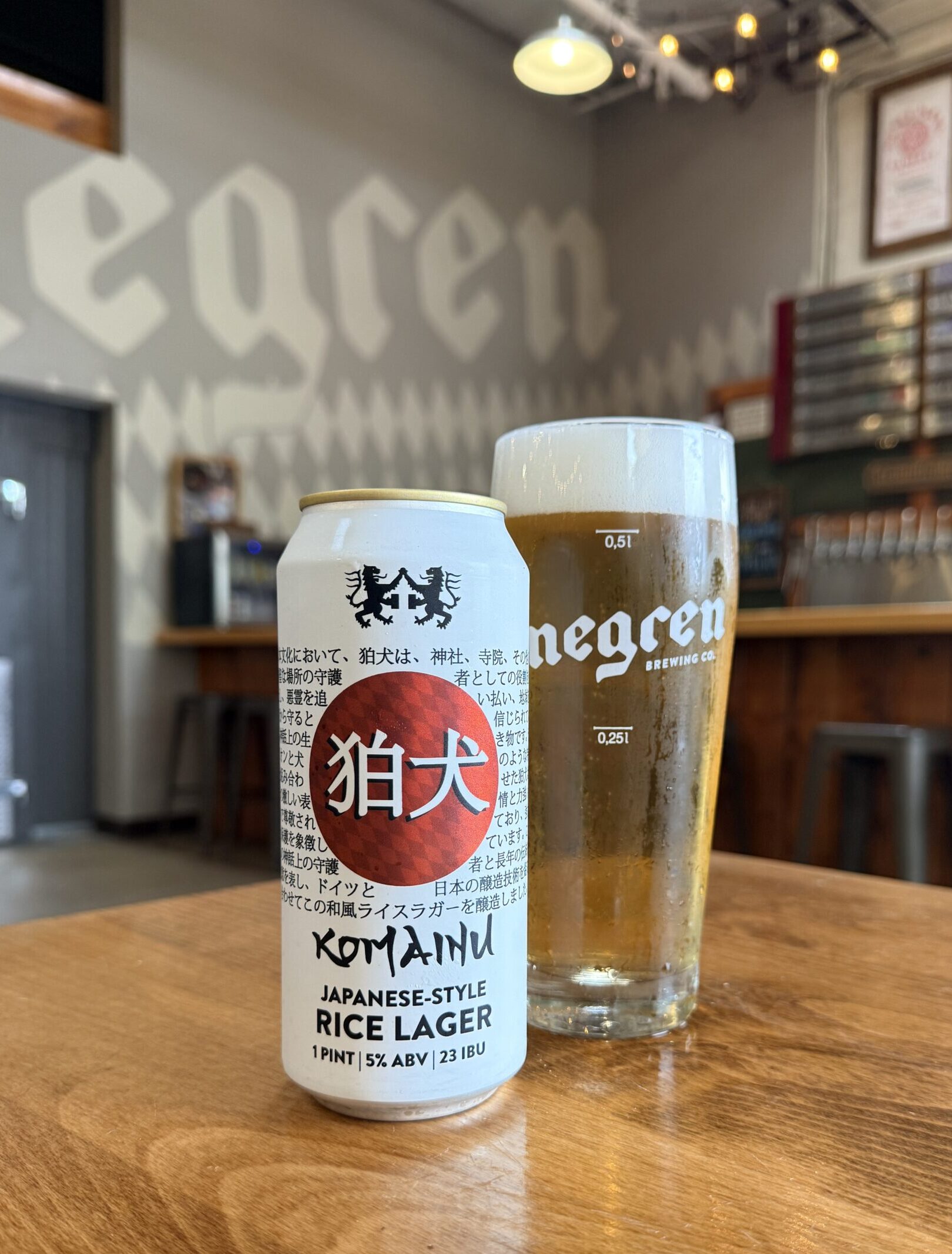Two beverages sit on a wooden table in a brewery: a white can labeled Komainu Japanese-style Rice Lager and a glass of beer nearby in the background.
